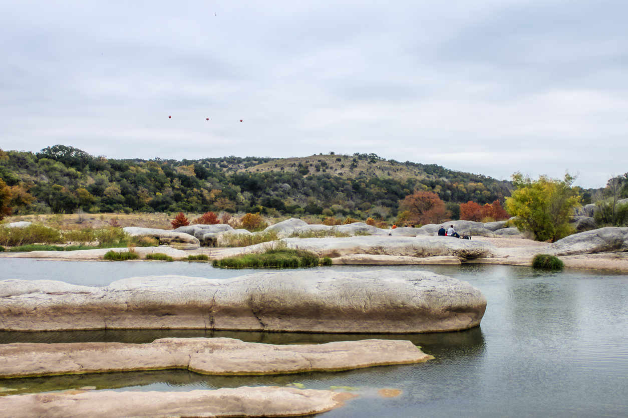 Large rocks in the water in the Pedernales River in the autumn in Texas with tourists sitting on rocks in distance and insulators on a high line above